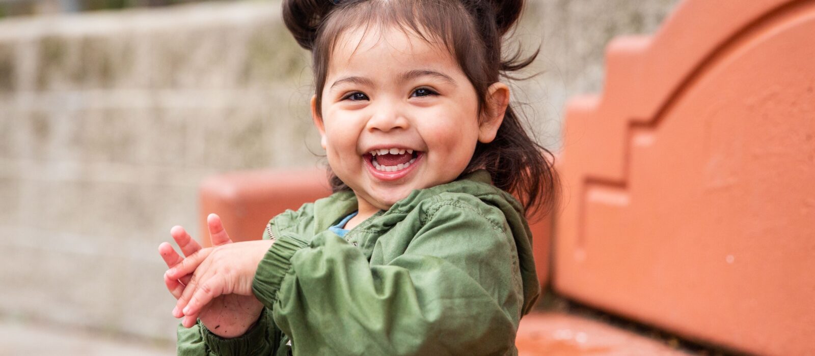 A toddler with medium skin tone sits on an outdoor bench, smiling joyfully.