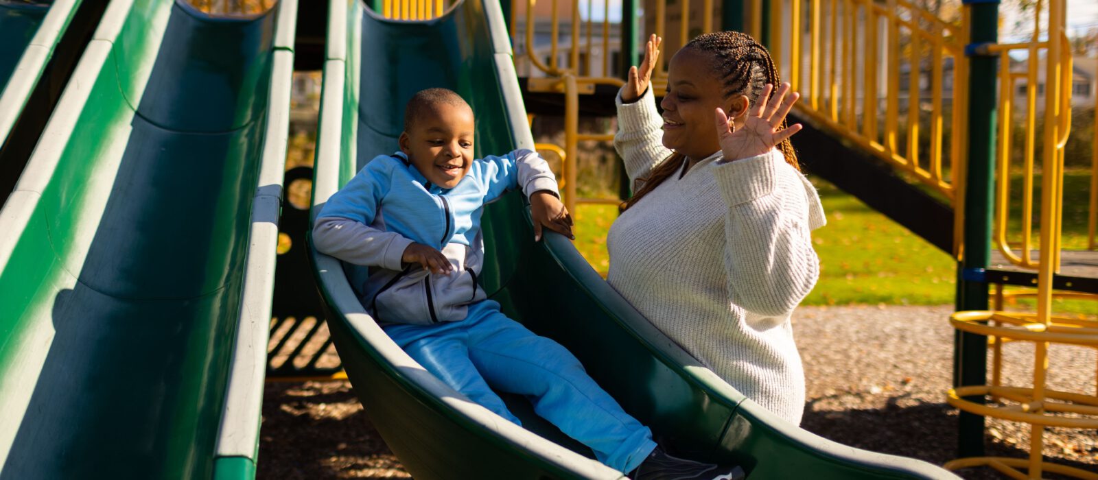 An adult cheering and smiling as their young child slides down a playground slide. Both have a dark skin tone.