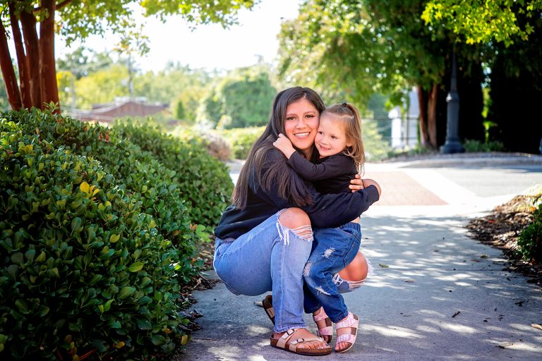An adult crouches on a sidewalk, hugging a toddler. Both have light skin tones and are smiling together, with trees and bushes in the background.