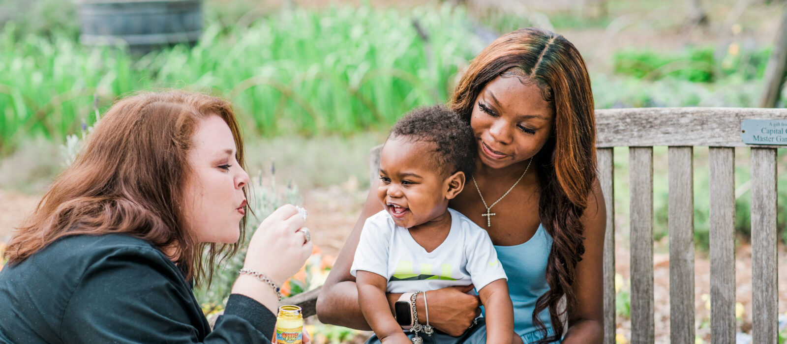 In a park, one adult with dark skin tone sits on a bench holding a baby with dark skin tone, while another adult with light skin tone blows bubbles, making the baby laugh.