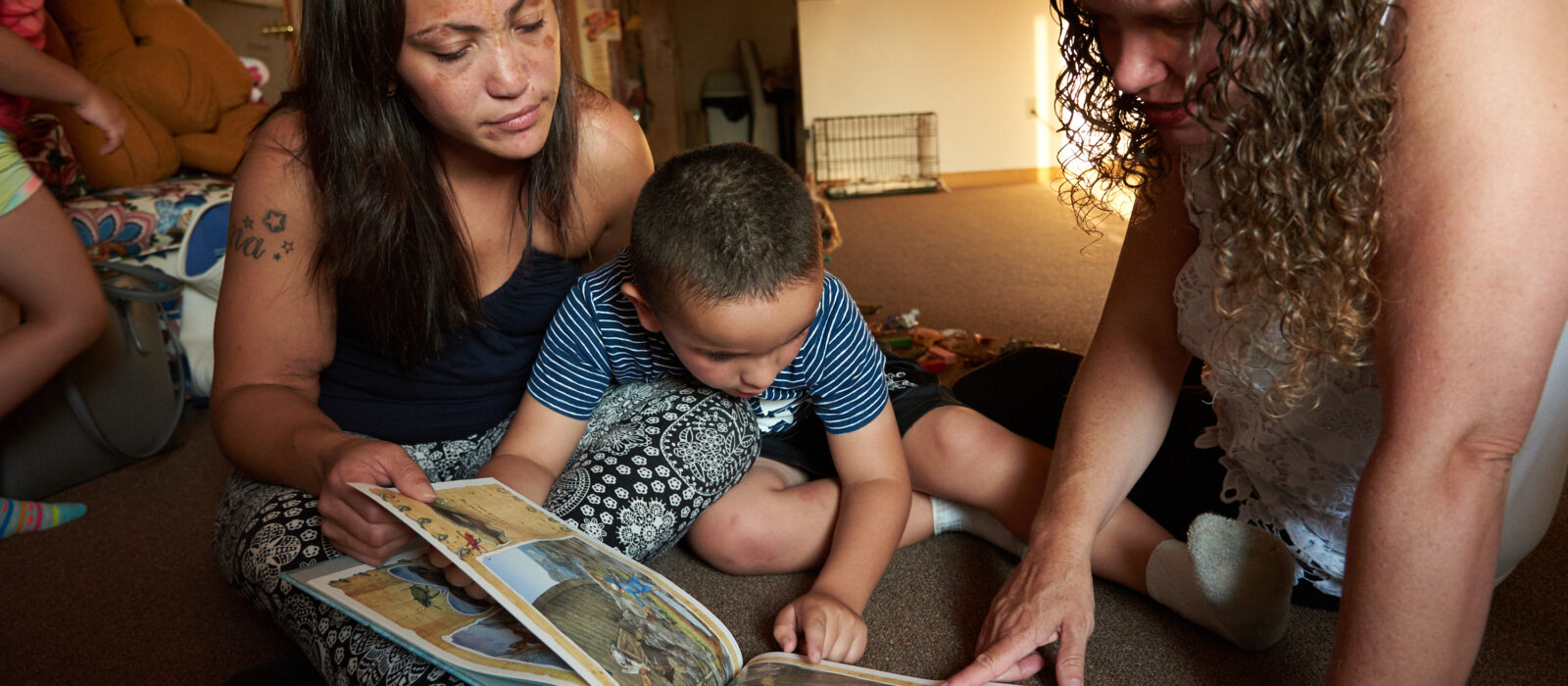 Two adults, one with medium skin tone and the other with light skin tone, sit on a living room floor, reading a book with a toddler who has medium skin tone.