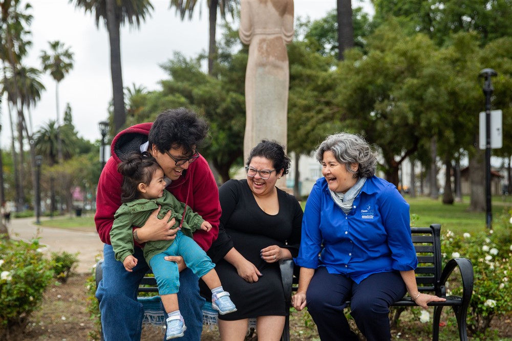 Two adults and one staff member from Nurse-Family Partnership, all with a medium skin tone, are sitting on a park bench with palm trees in the background, laughing together with a baby who has a medium skin tone on the adult's lap.