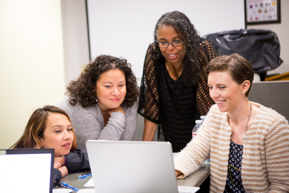 Four adults of various skin tones gather together looking at a computer