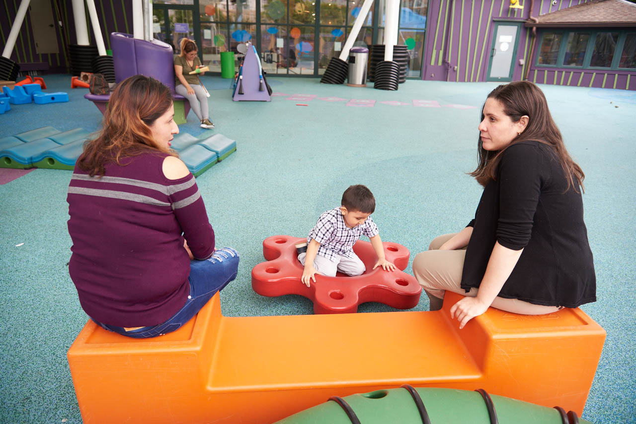 Two adults, both with a medium skin tone, are sitting on a bench in an indoor playground while a child with a medium skin tone plays nearby.