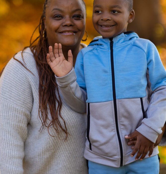 An adult with a dark skin tone stands in a park, smiling while posing with a child who also has a dark skin tone.