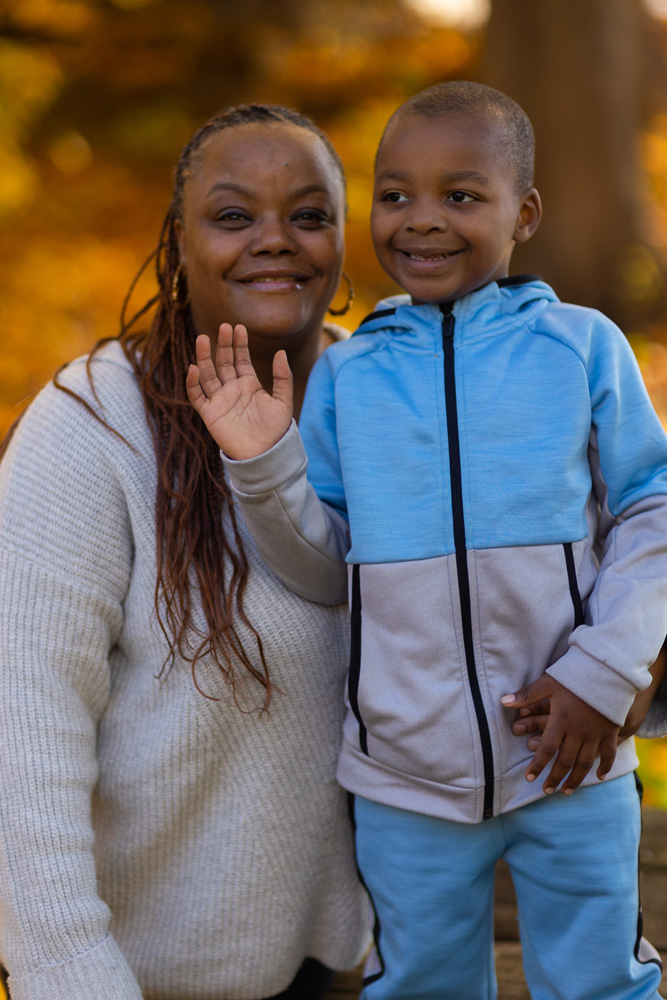 An adult with a dark skin tone stands in a park, smiling while posing with a child who also has a dark skin tone.