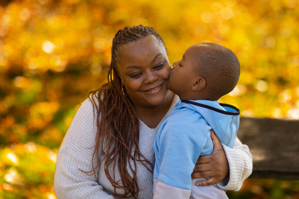 A young child is kissing an adult on the cheek outdoors, both with a dark skin tone, with fall leaves in the background.