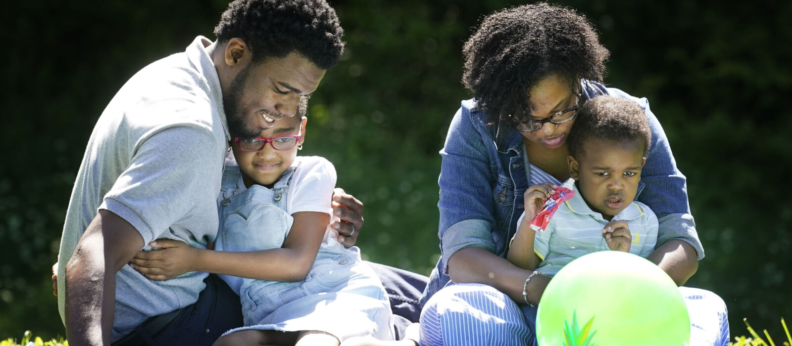 A family of four with a dark skin tone is sitting on a blanket in the park, with two children in the adults’ laps, all focused on a ball in front of them.