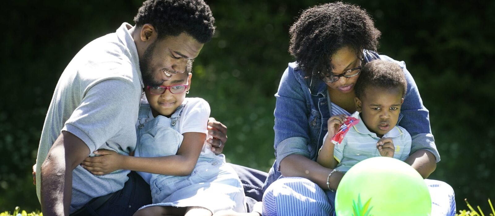 A family of four with a dark skin tone is sitting on a blanket in the park, with two children in the adults’ laps, all focused on a ball in front of them.