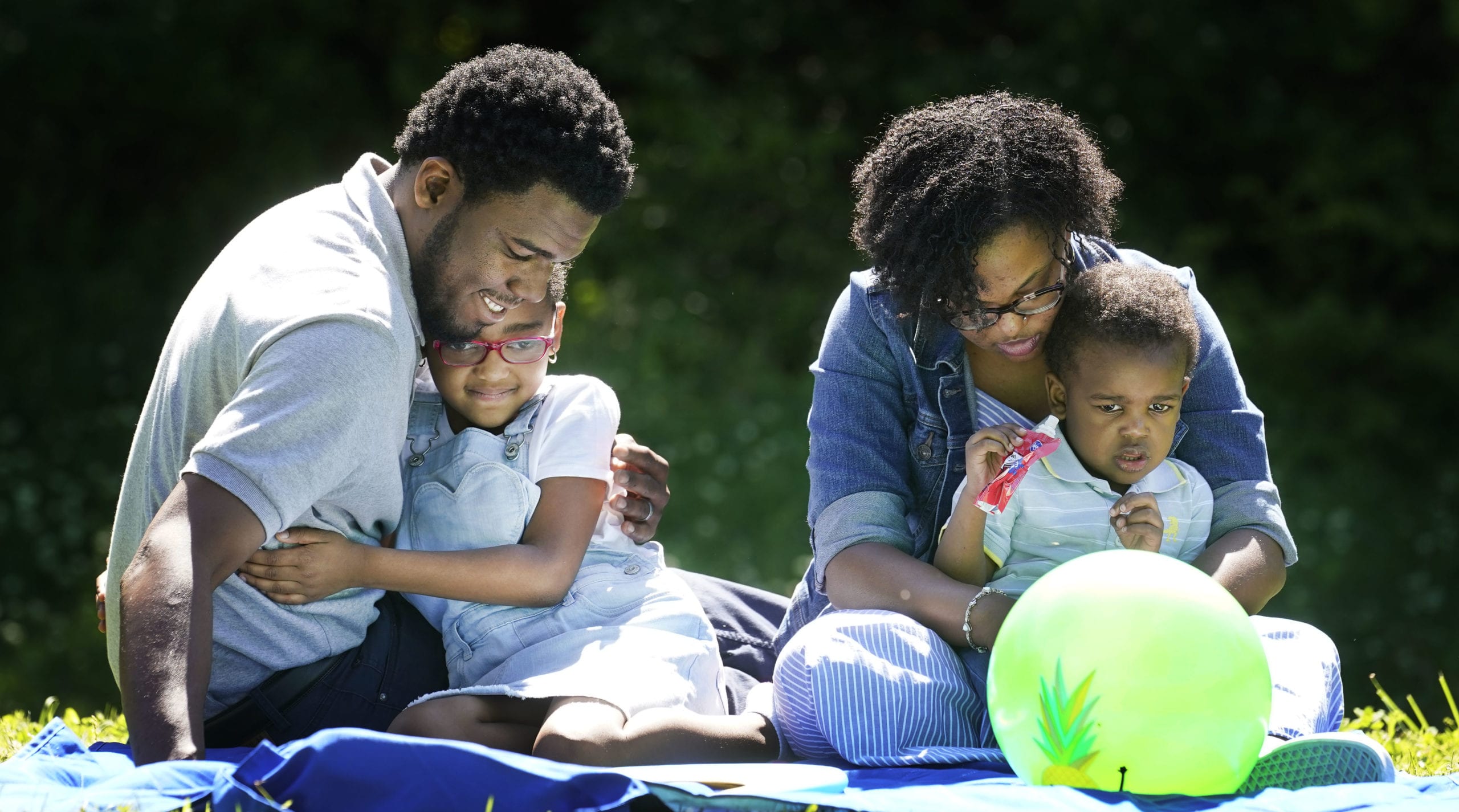 A family of four with a dark skin tone is sitting on a blanket in the park, with two children in the adults’ laps, all focused on a ball in front of them.
