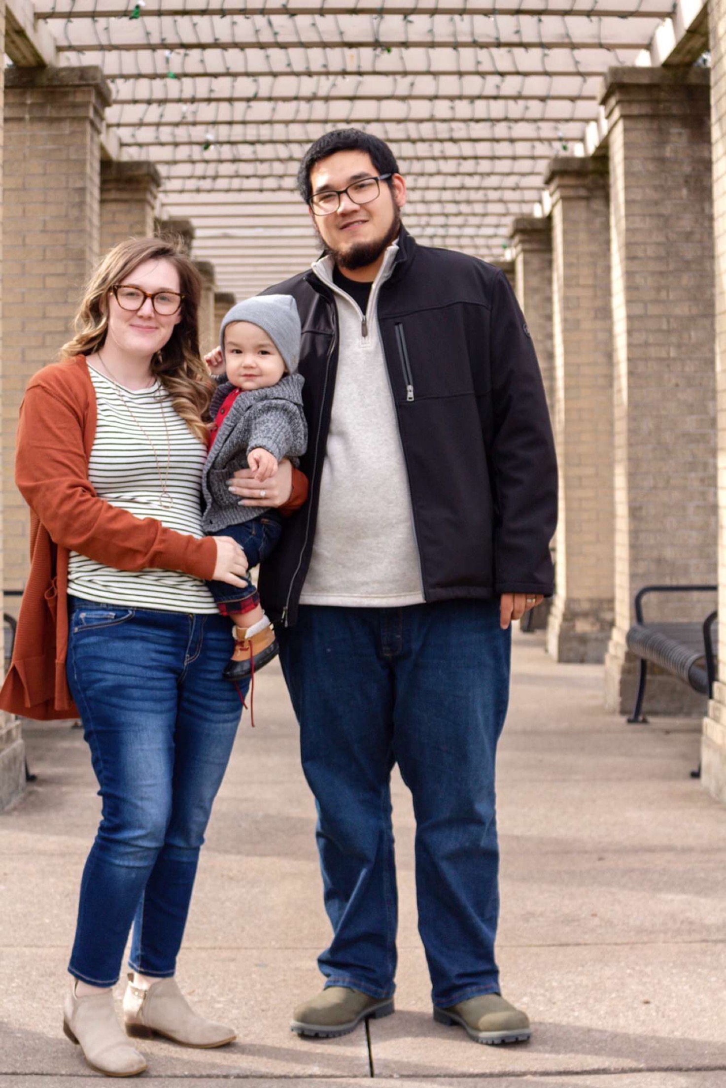 Two adults, one with a medium skin tone and the other with a light skin tone, stand under a pavilion while the adult with a light skin tone holds a baby with a light skin tone.
