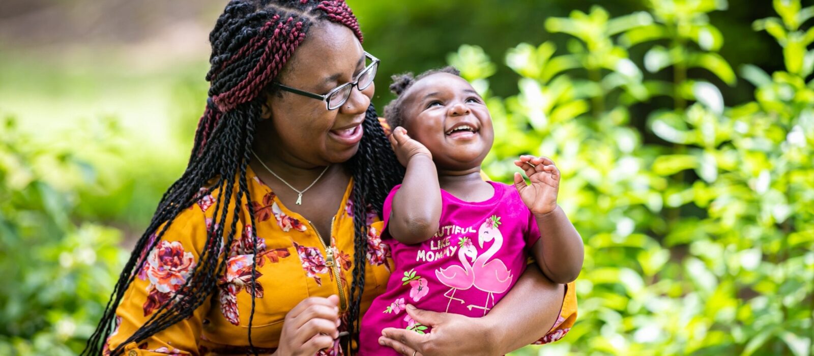 An adult with a dark skin tone is holding a child with a dark skin tone in their lap in front of lush greenery. The adult looks at the child, who is looking up and laughing.