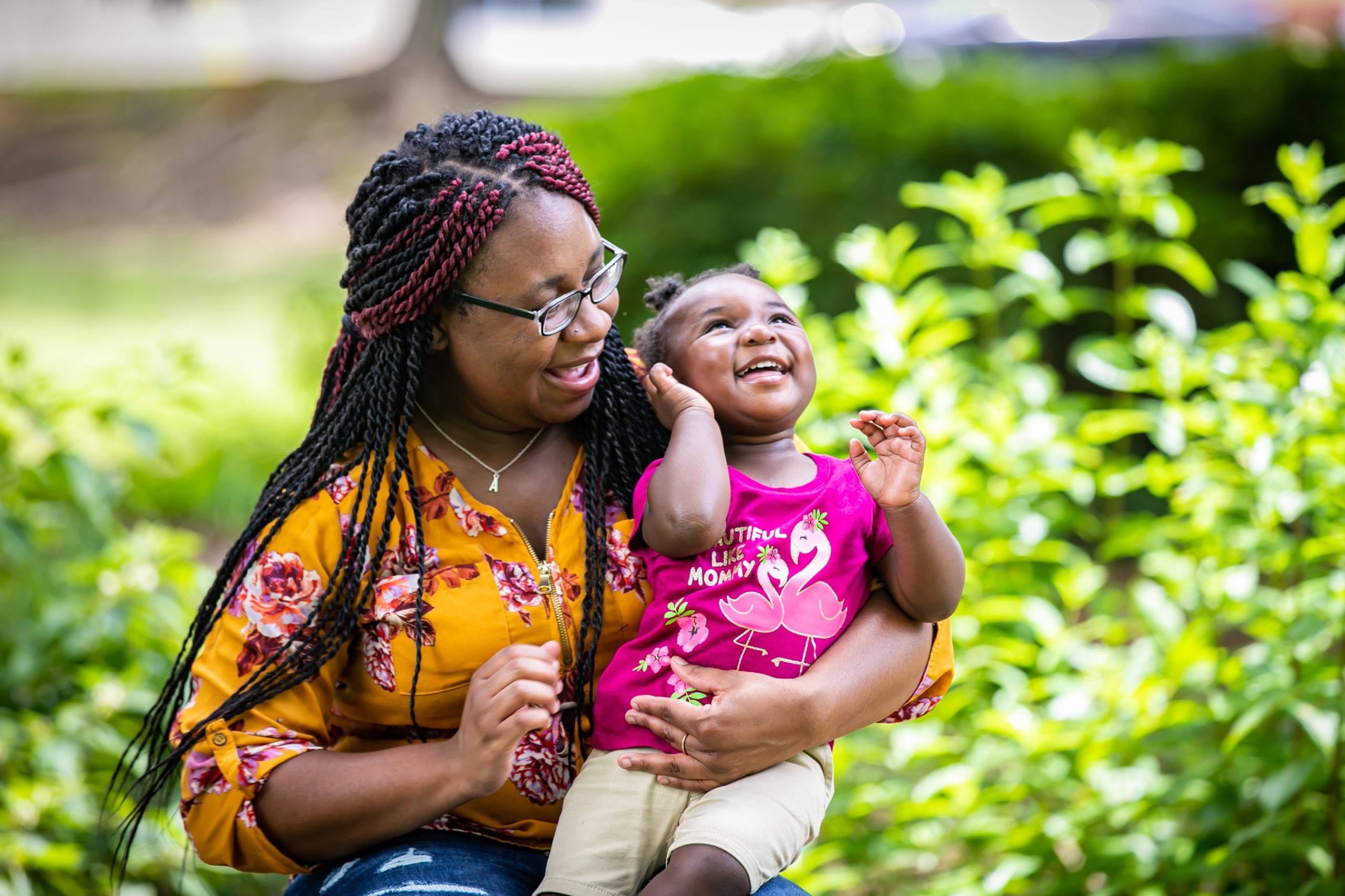 An adult with a dark skin tone is holding a child with a dark skin tone in their lap in front of lush greenery. The adult looks at the child, who is looking up and laughing.
