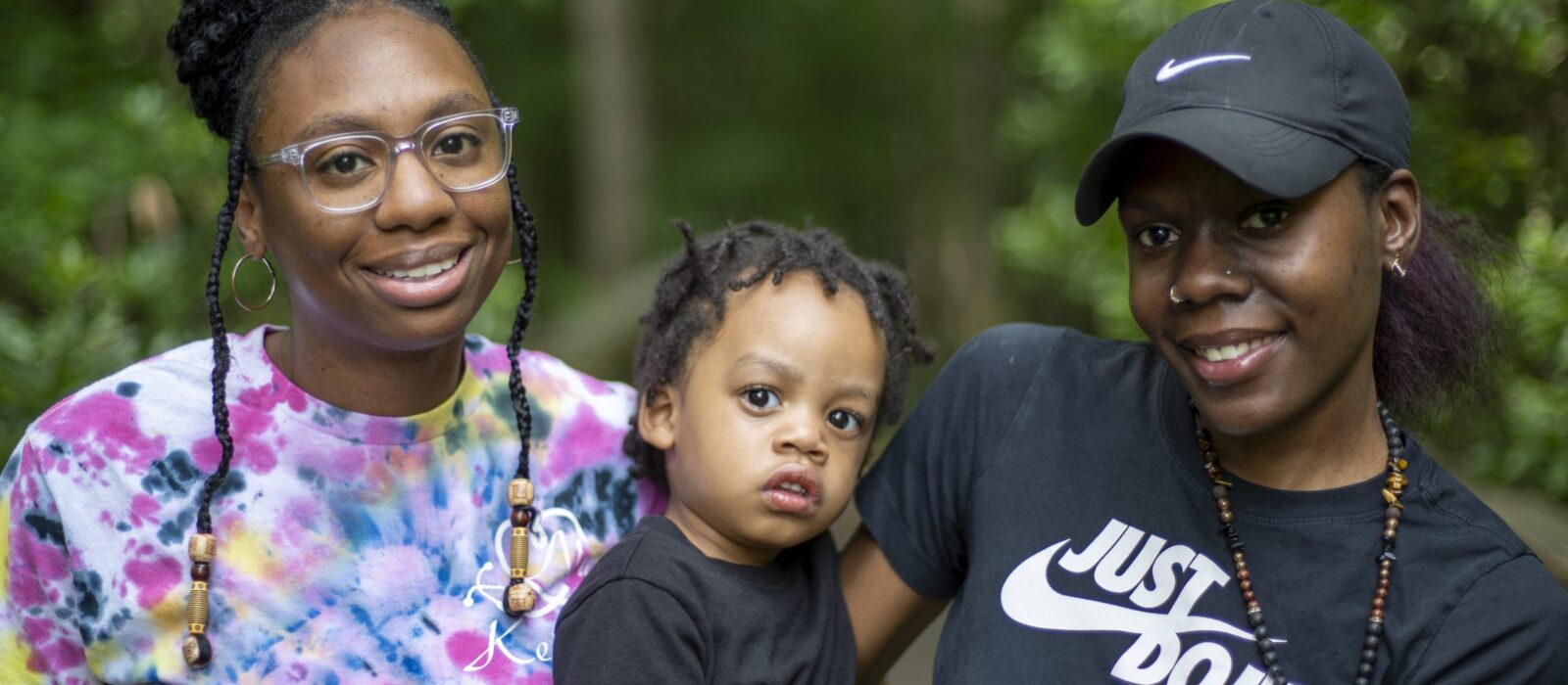 An adult and a teenager, both with a dark skin tone, are standing in front of trees, with a baby who has a dark skin tone between them.