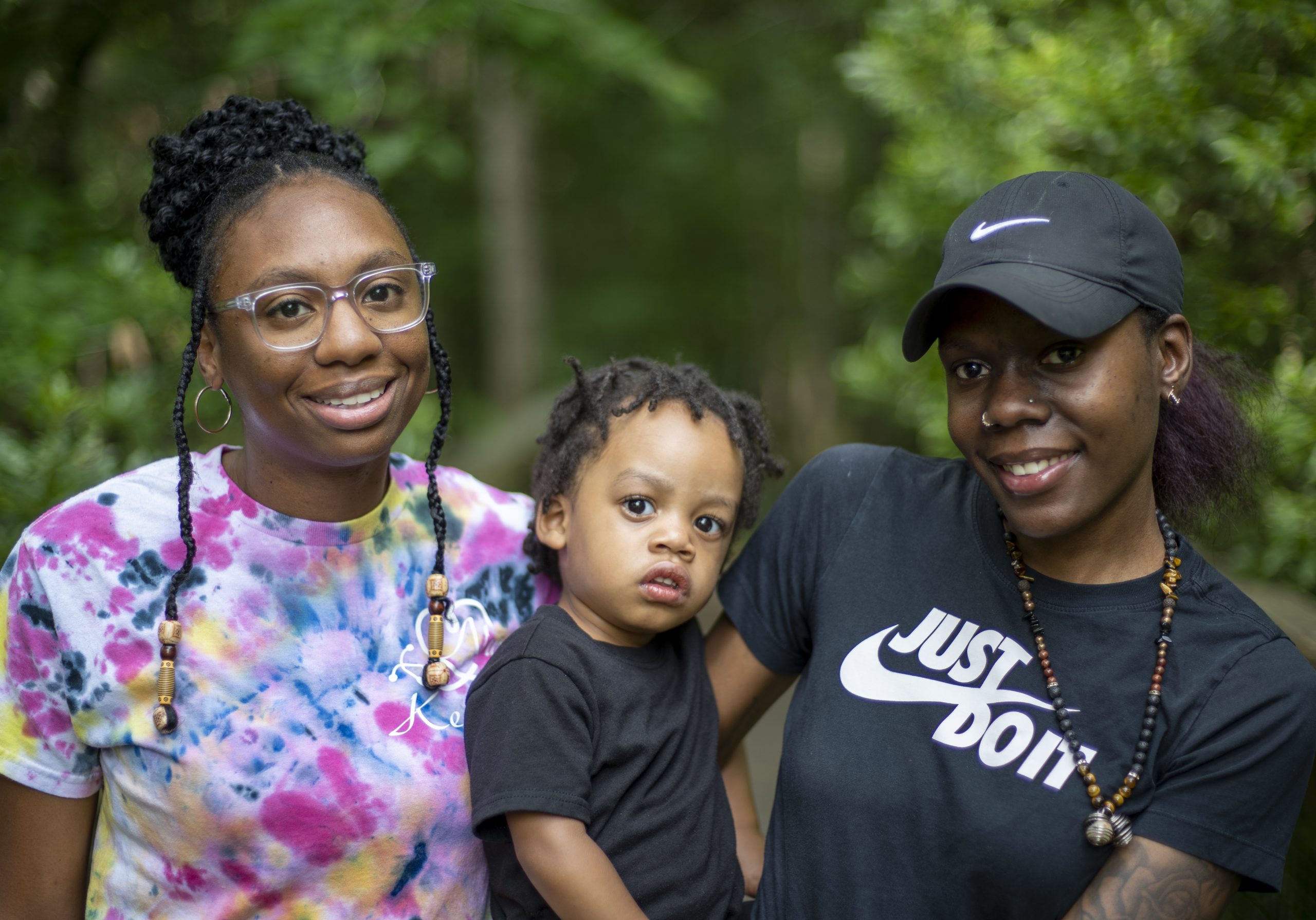 An adult and a teenager, both with a dark skin tone, are standing in front of trees, with a baby who has a dark skin tone between them.