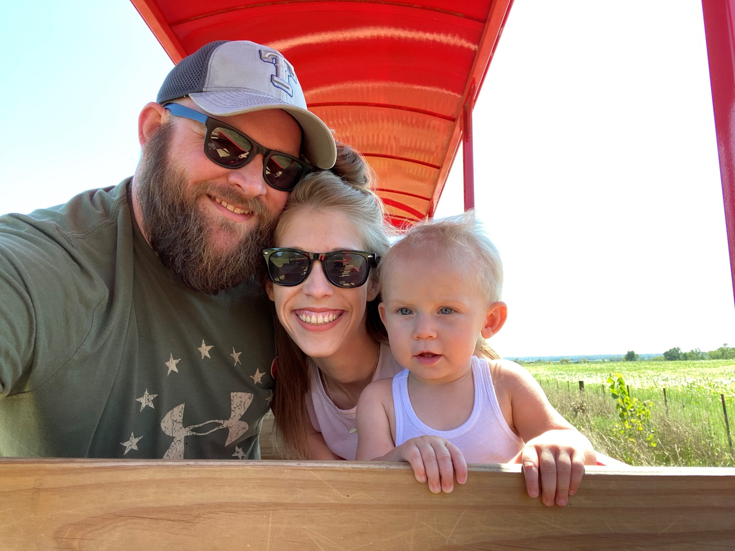 Two adults with a light skin tone are sitting on a wagon with a baby who has a light skin tone in their lap, with fields visible behind them.