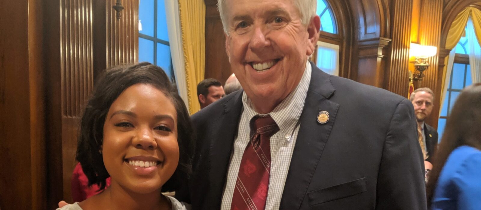 An adult with a light skin tone and an adult with a dark skin tone are standing side by side, smiling for a photo at an event.