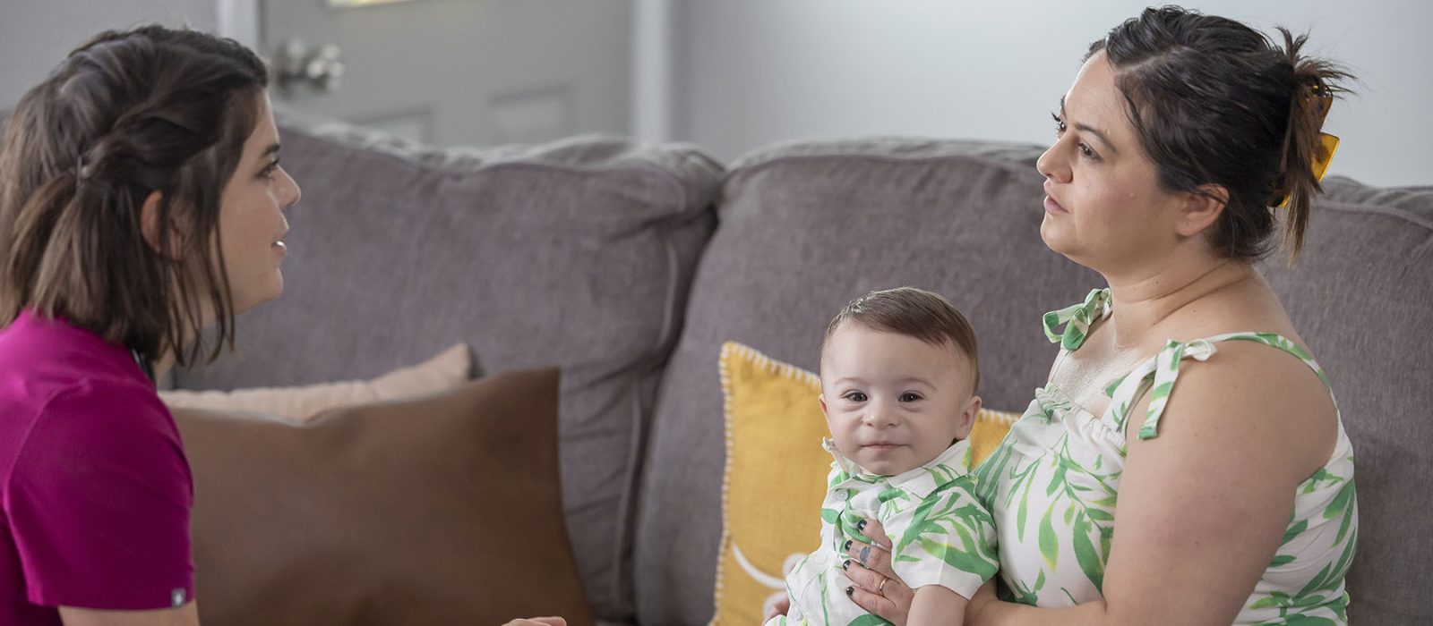 A female nurse with a light skin tone and wearing scrubs is sitting with a mom and her baby, both with a light skin tone, in a living room, engaged in a conversation.