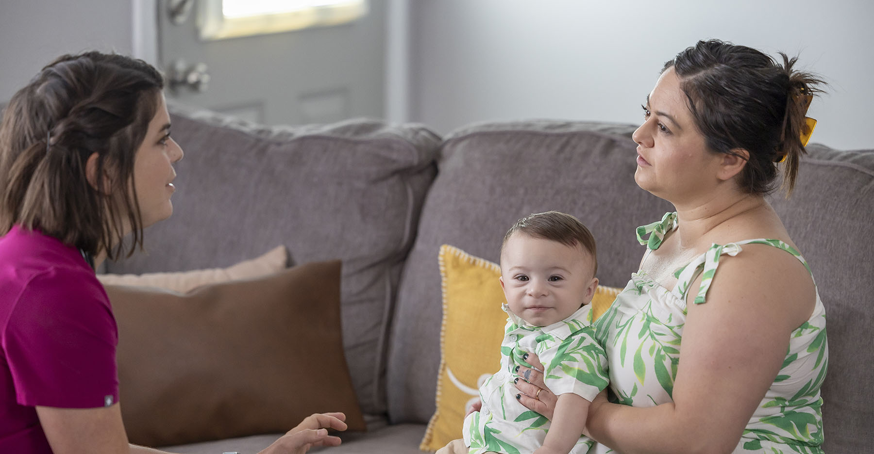 A female nurse with a light skin tone and wearing scrubs is sitting with a mom and her baby, both with a light skin tone, in a living room, engaged in a conversation.
