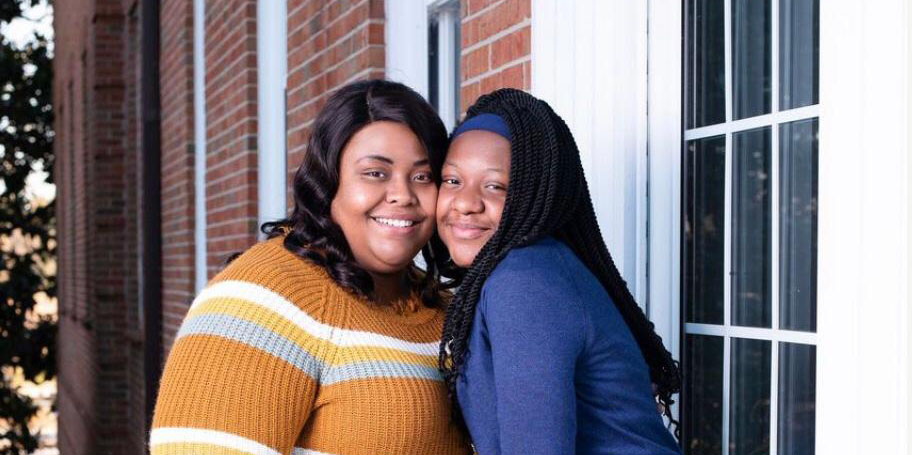 An adult and a teenager, both with a dark skin tone, are standing together in front of a brick building with windows, smiling for a photo.