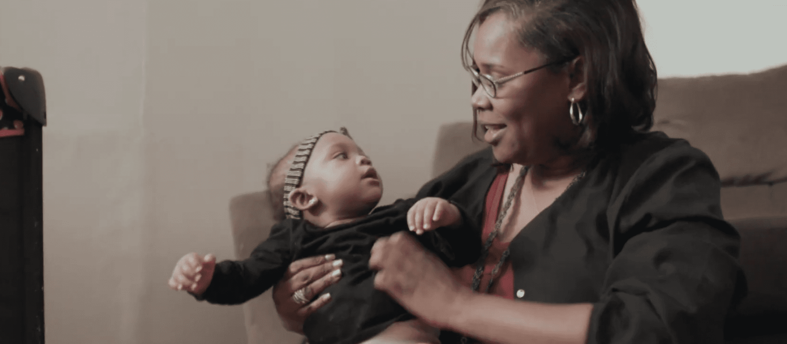 An adult with a dark skin tone is sitting in a cozy living room, smiling warmly while holding a baby with a dark skin tone.