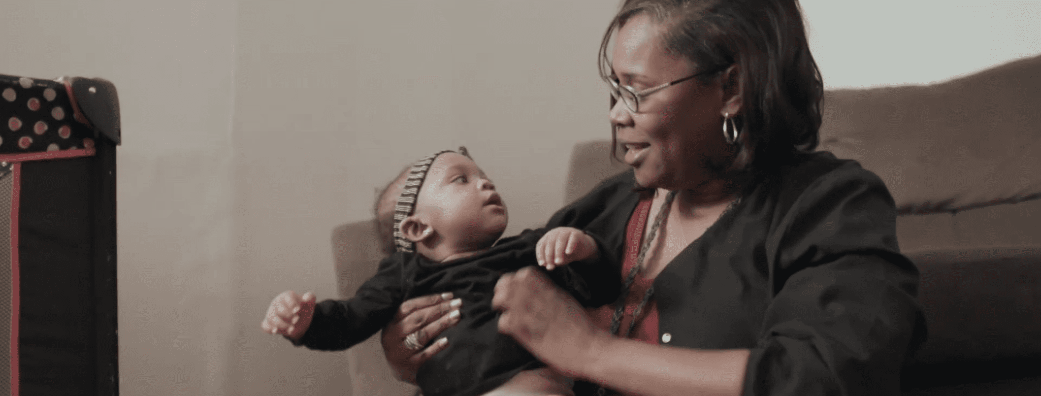 An adult with a dark skin tone is sitting in a cozy living room, smiling warmly while holding a baby with a dark skin tone.