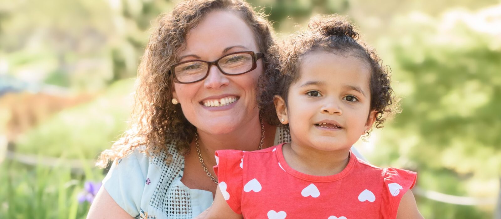 An adult with a medium skin tone is sitting in a park with a toddler with a medium skin tone, surrounded by lush greenery in the background.