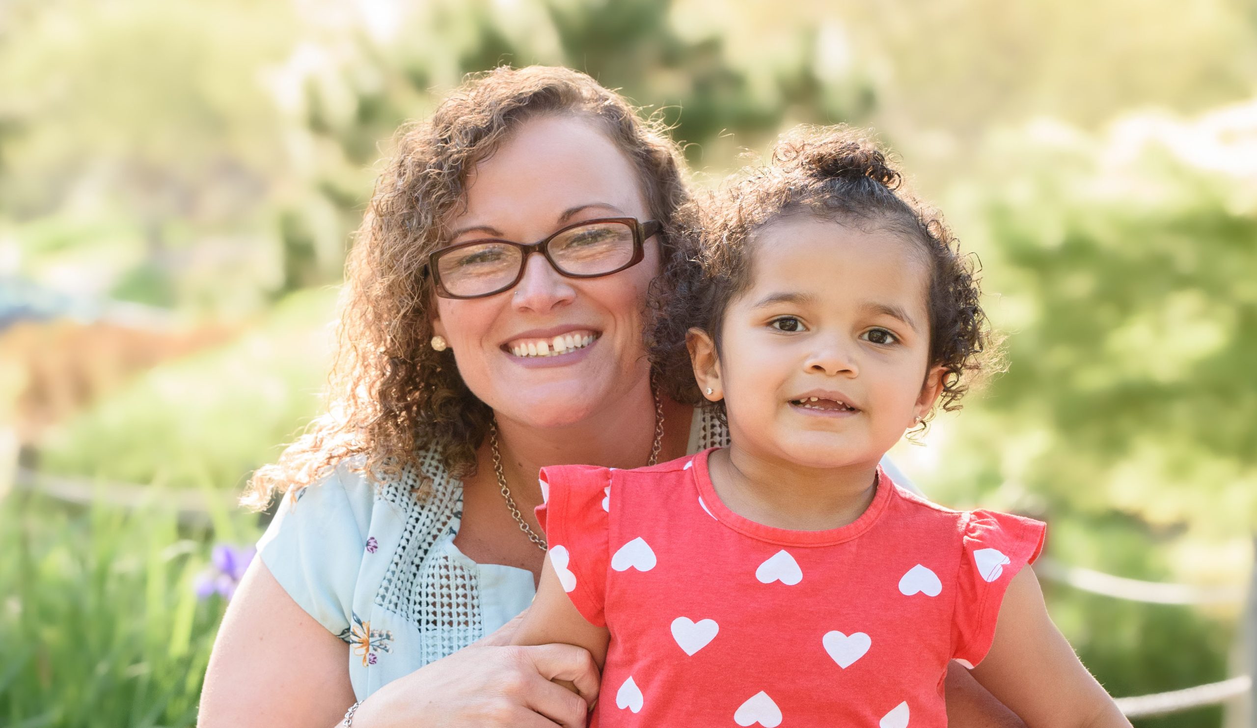 An adult with a medium skin tone is sitting in a park with a toddler with a medium skin tone, surrounded by lush greenery in the background.