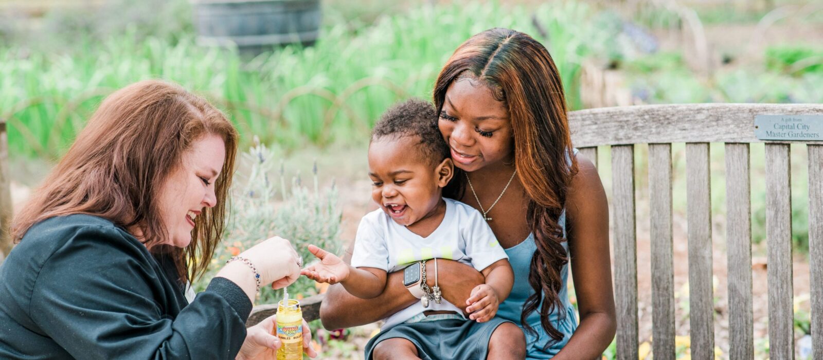 Two adults, one with a dark skin tone and one with a light skin tone, are in a park. One adult, with a dark skin tone, is sitting on a bench holding a baby, while the other, with a light skin tone, blows bubbles to make the baby laugh.