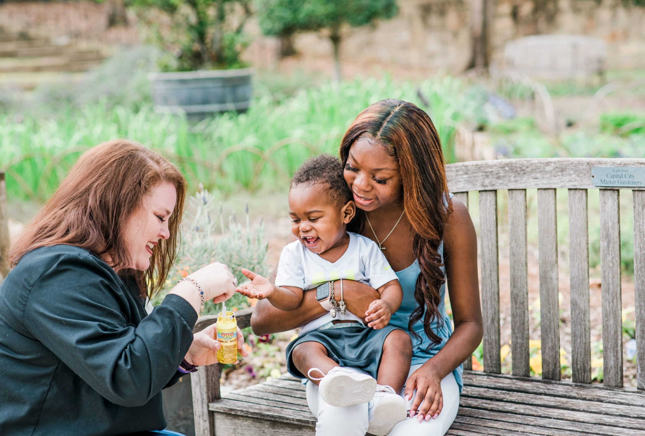 Two adults, one with a dark skin tone and one with a light skin tone, are in a park. One adult, with a dark skin tone, is sitting on a bench holding a baby, while the other, with a light skin tone, blows bubbles to make the baby laugh.