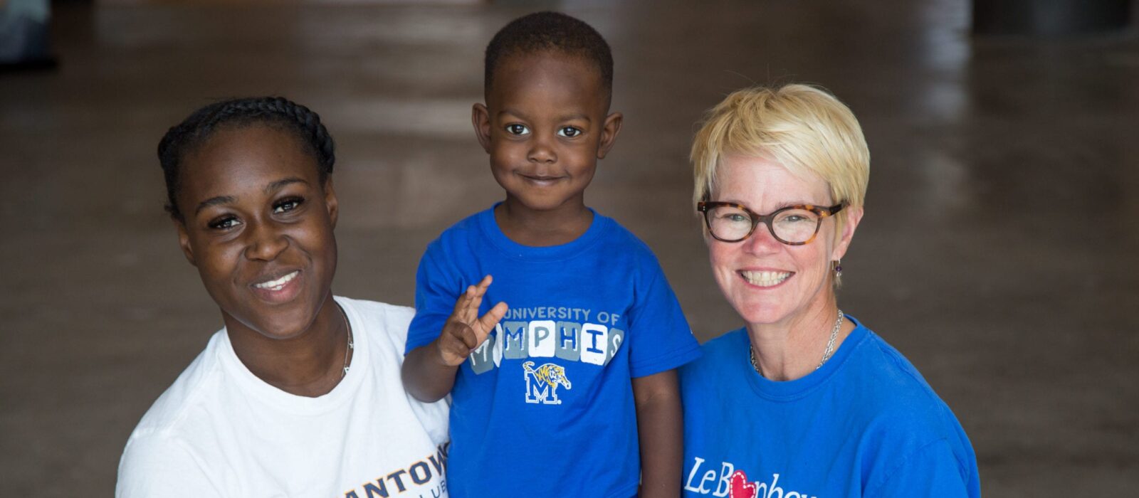 Two adults, one with a light skin tone and one with a dark skin tone, are standing in a dimly lit room, smiling with a toddler between them who has a dark skin tone.