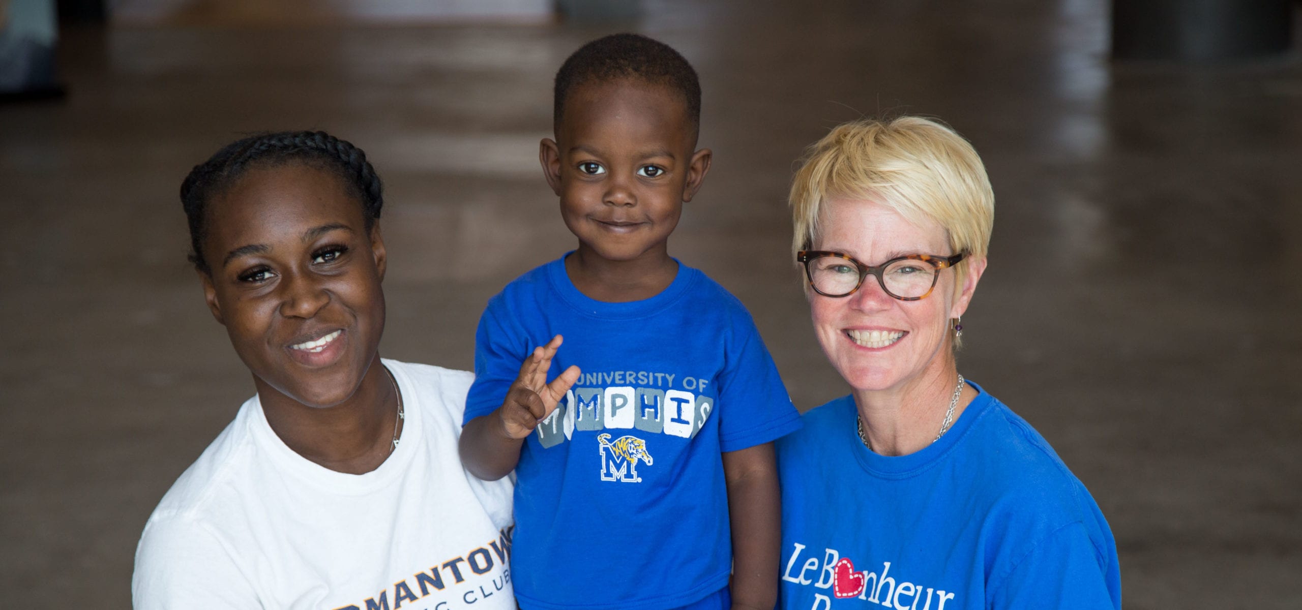 Two adults, one with a light skin tone and one with a dark skin tone, are standing in a dimly lit room, smiling with a toddler between them who has a dark skin tone.