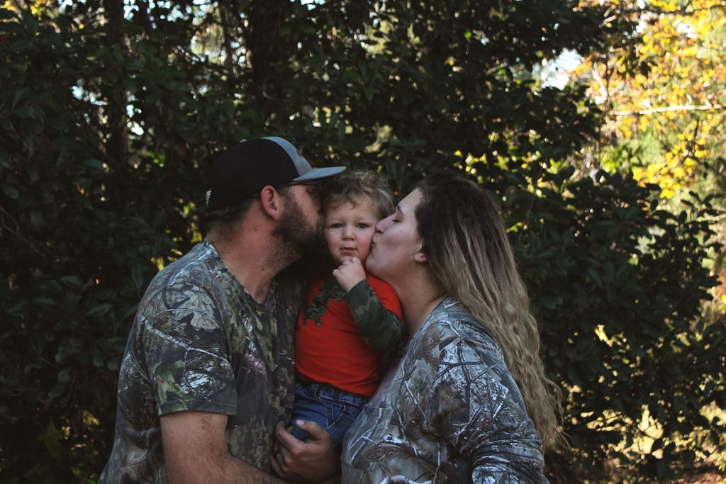 An adult with a light skin tone and an adult with a light skin tone are kissing a toddler with a light skin tone on the cheeks while posing in front of trees.