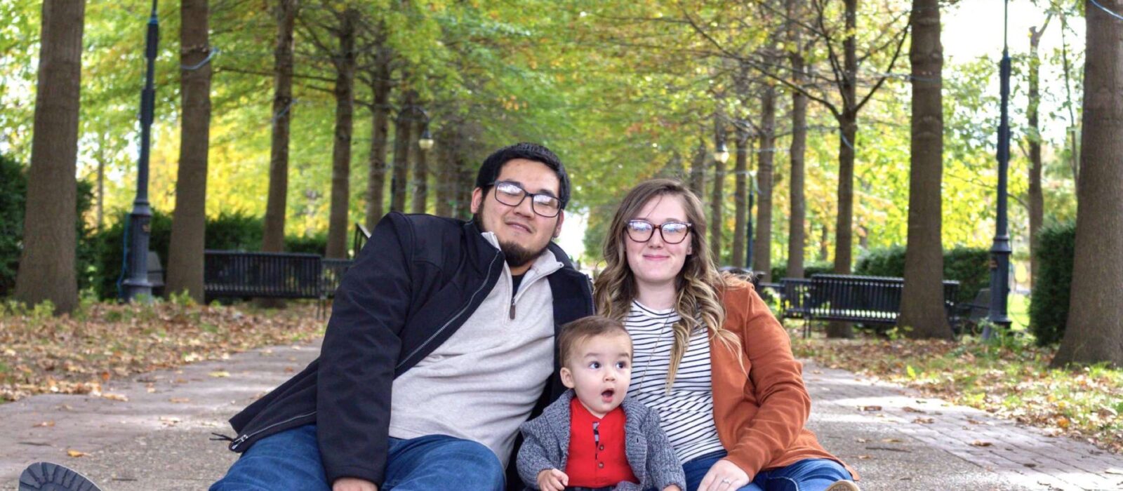 An adult with a medium skin tone and an adult with a light skin tone are sitting on a sidewalk in a shaded tree grove with a baby who has a light skin tone between them.