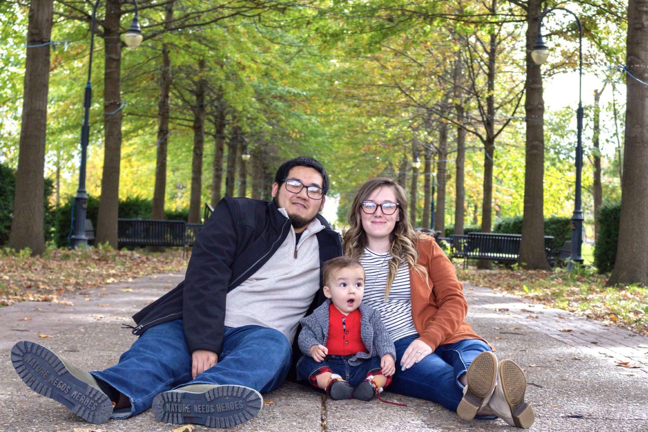 An adult with a medium skin tone and an adult with a light skin tone are sitting on a sidewalk in a shaded tree grove with a baby who has a light skin tone between them.