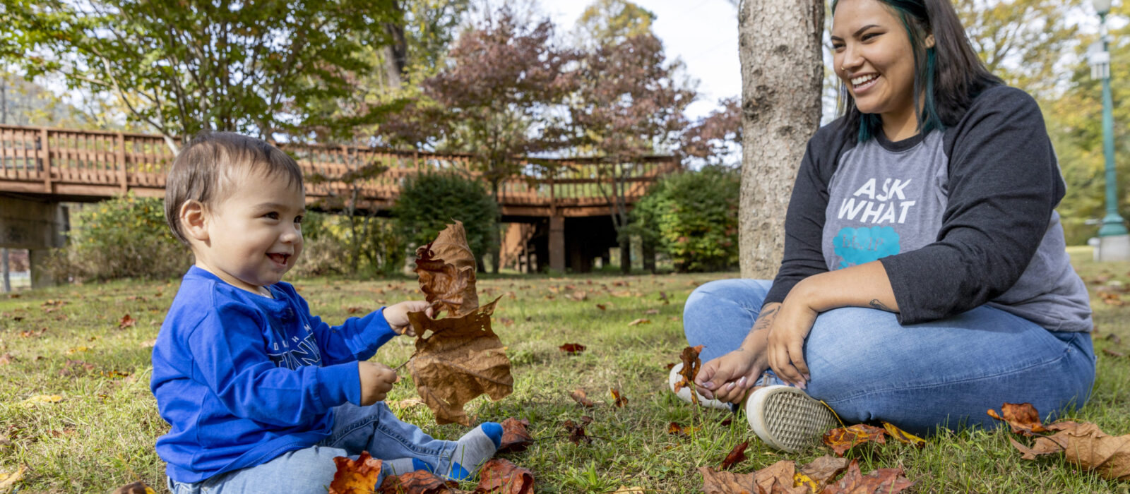 An adult with a medium skin tone is sitting on the grass with a smiling baby who has a medium skin tone, playing together with fallen leaves.