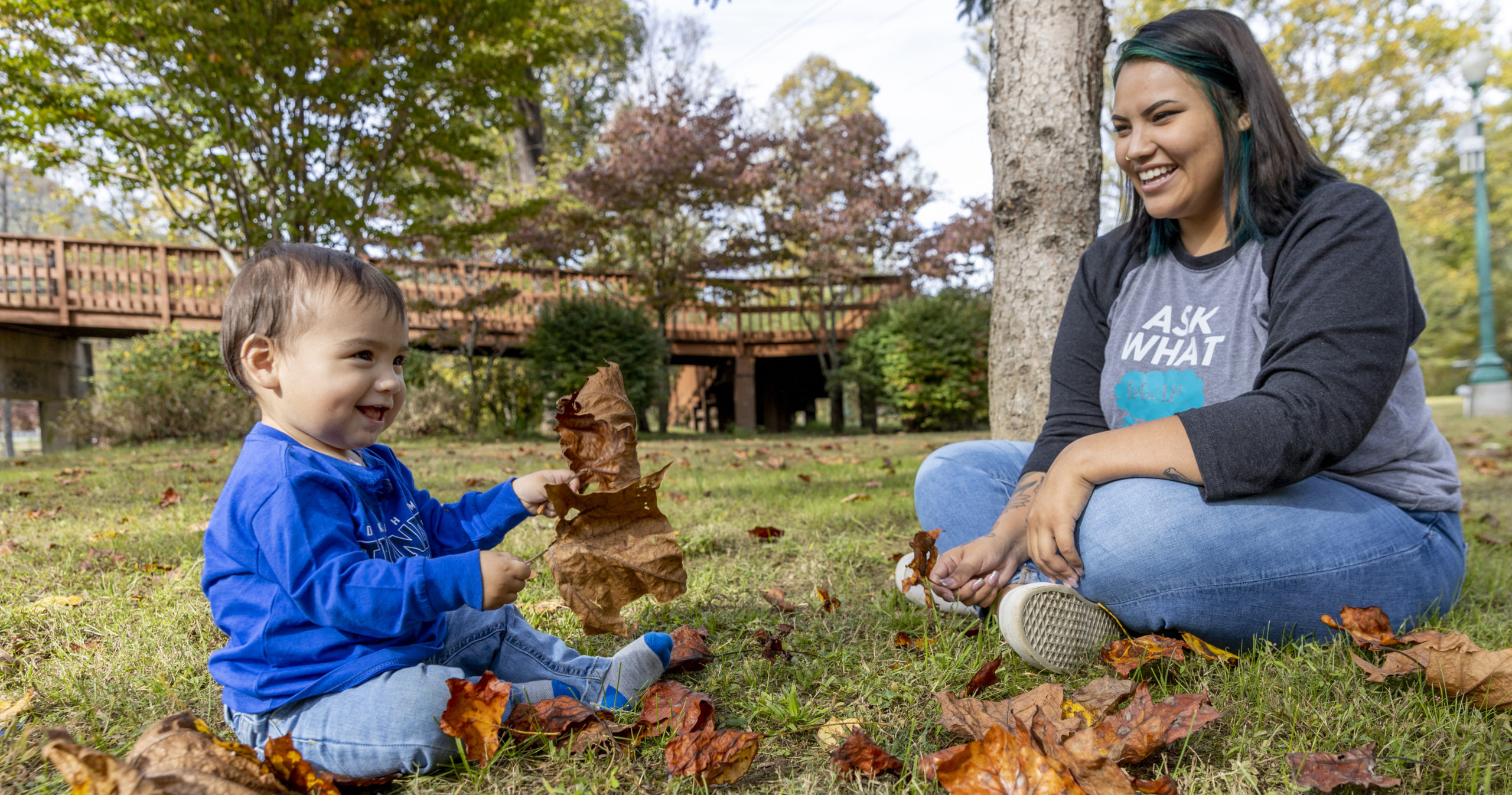 An adult with a medium skin tone is sitting on the grass with a smiling baby who has a medium skin tone, playing together with fallen leaves.