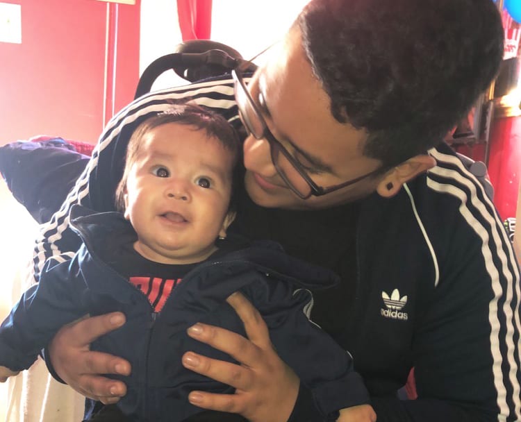 An adult with a medium skin tone is sitting on a living room floor, smiling warmly as they look down at a baby with a medium skin tone.