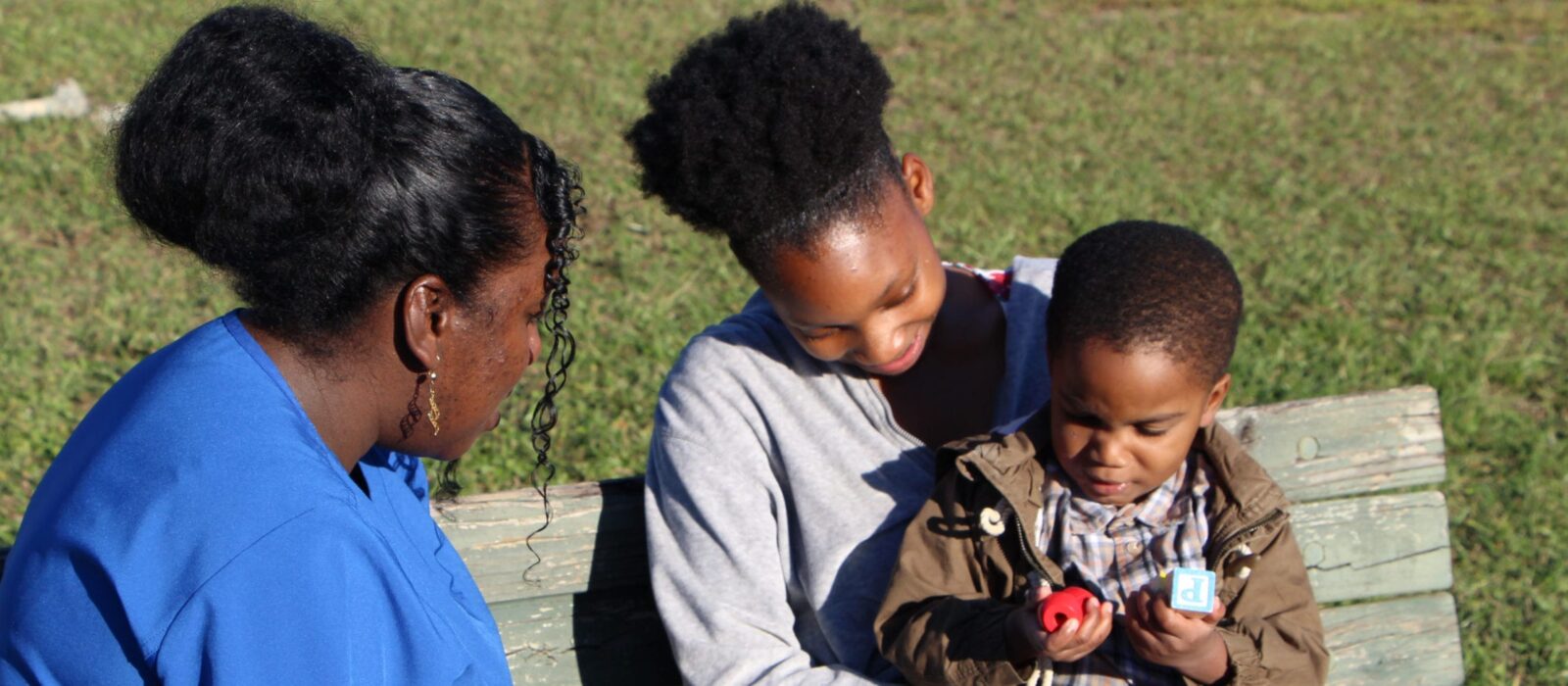 An adult with a dark skin tone and a baby are sitting on a park bench with a nurse, also with a dark skin tone. They are watching as the baby on the adult’s lap plays with a toy.