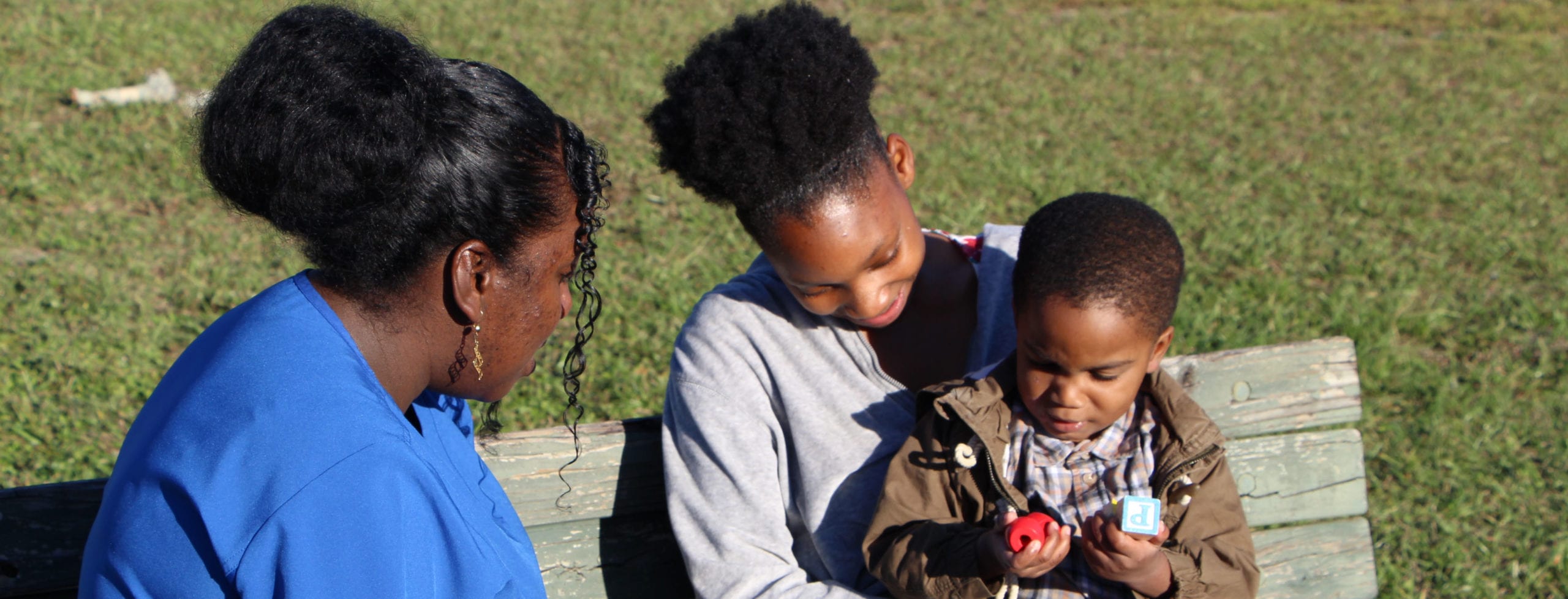 An adult with a dark skin tone and a baby are sitting on a park bench with a nurse, also with a dark skin tone. They are watching as the baby on the adult’s lap plays with a toy.