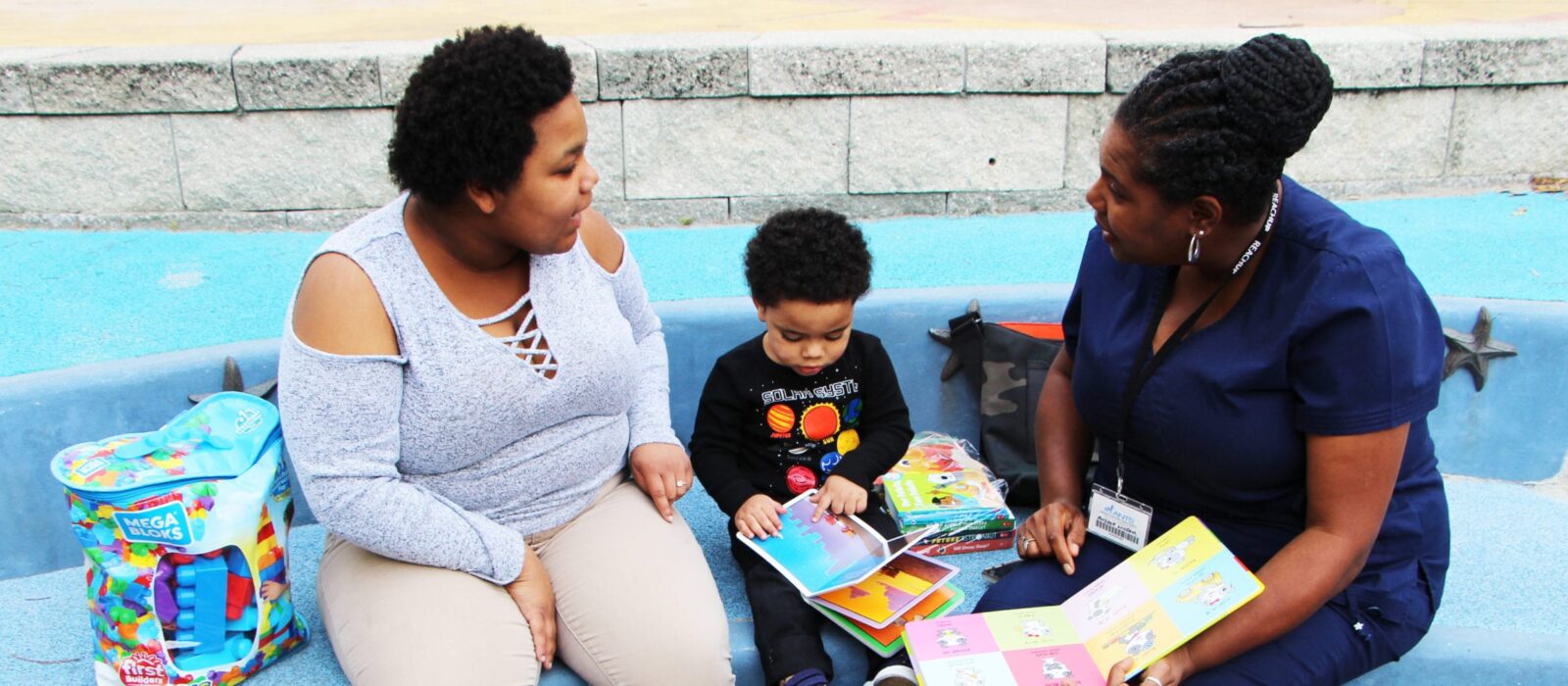 An adult and a toddler, both with a dark skin tone, are sitting in a park with a nurse who also has a dark skin tone. The nurse is reading a book to them during a visit.