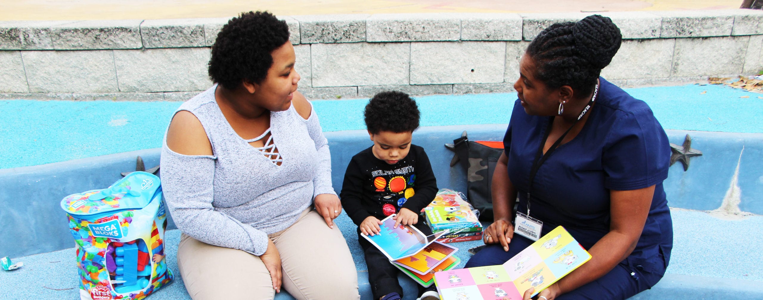 An adult and a toddler, both with a dark skin tone, are sitting in a park with a nurse who also has a dark skin tone. The nurse is reading a book to them during a visit.