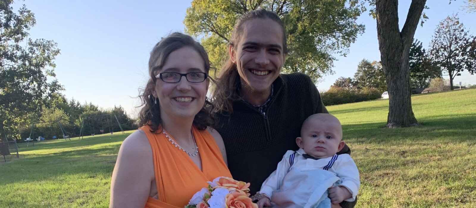 Two adults with a light skin tone are standing together in front of trees, posing with their child who has a light skin tone.