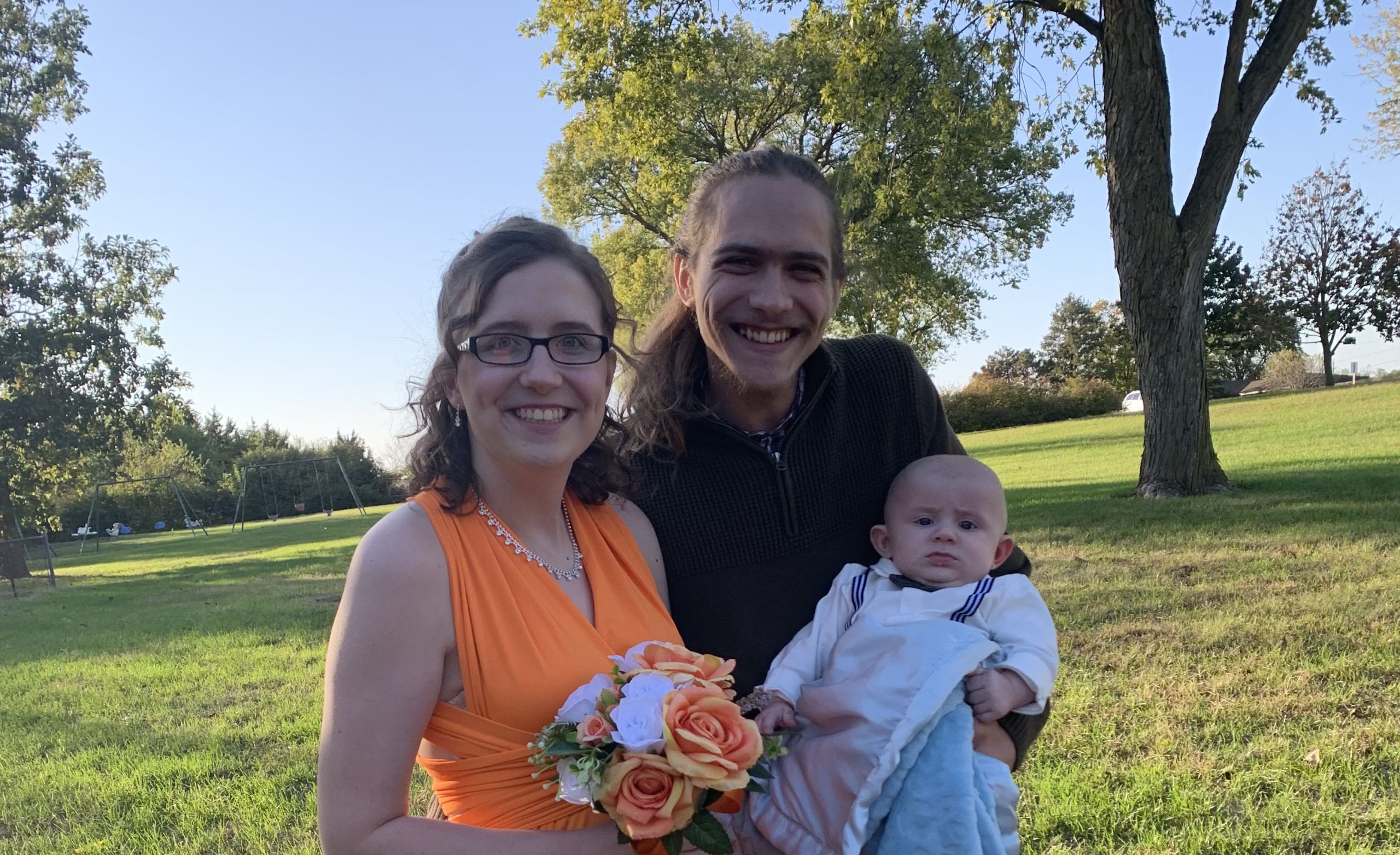 Two adults with a light skin tone are standing together in front of trees, posing with their child who has a light skin tone.