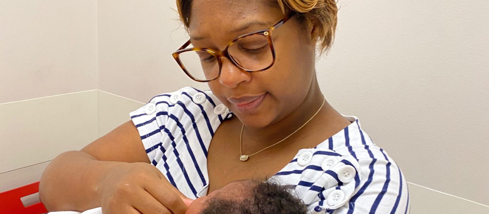An adult with a dark skin tone is sitting indoors, gently touching their baby’s face as they look down at them in their arms.
