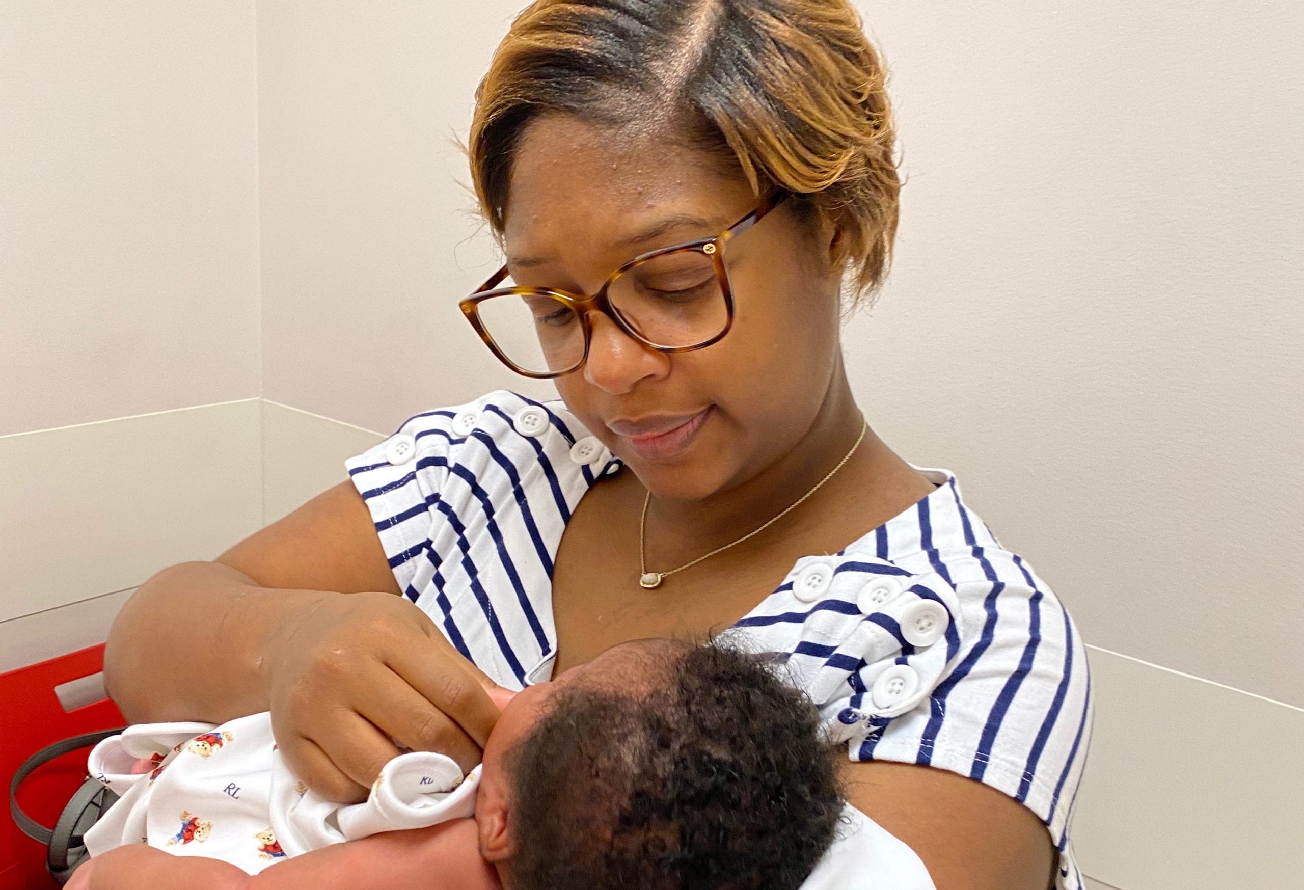 An adult with a dark skin tone is sitting indoors, gently touching their baby’s face as they look down at them in their arms.