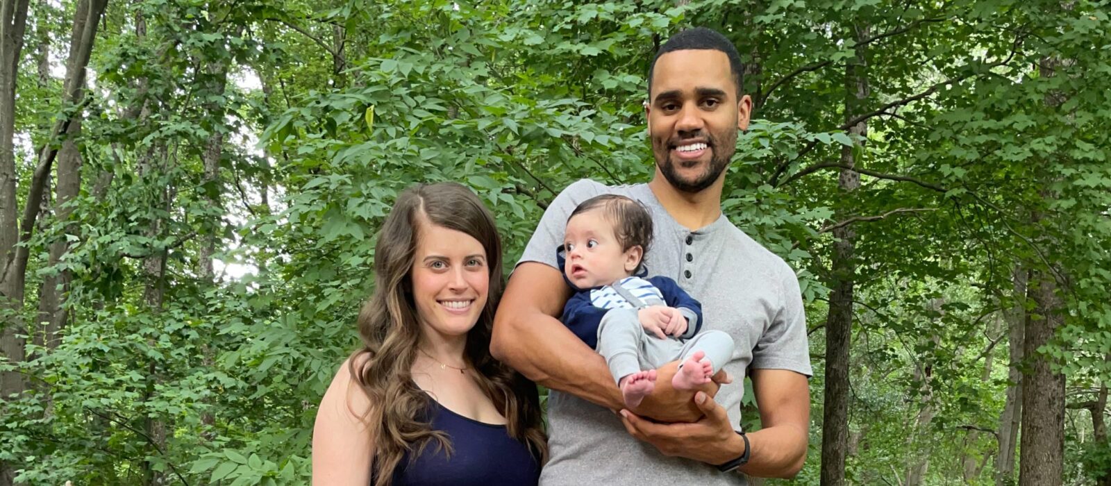 Two adults, one with a light skin tone and one with a medium skin tone, are standing together in front of trees, posing with their child who has a medium skin tone.