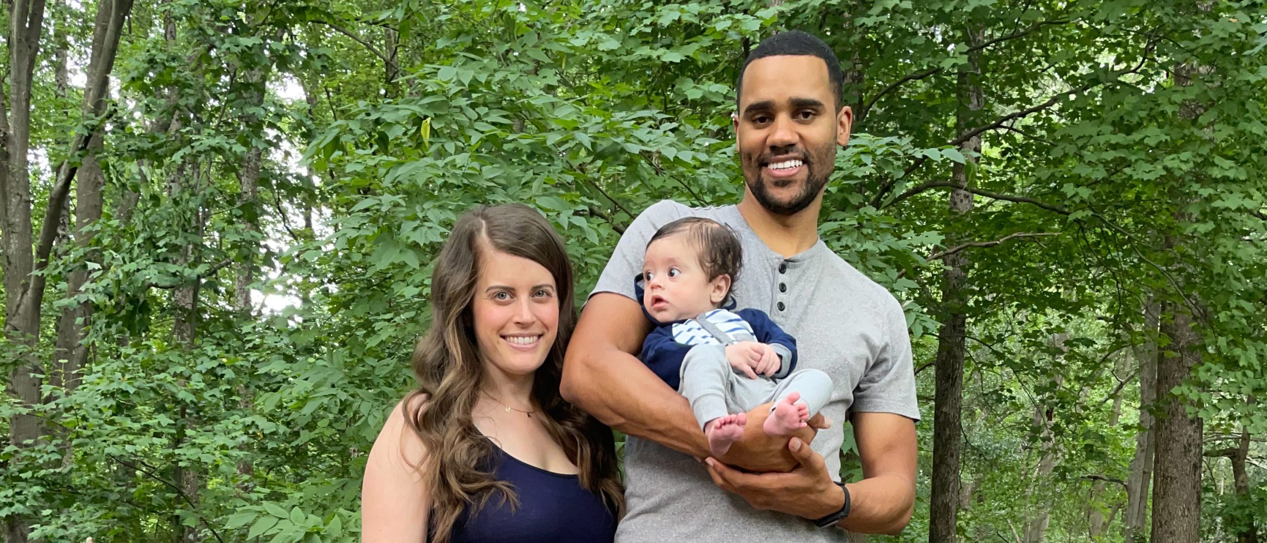 Two adults, one with a light skin tone and one with a medium skin tone, are standing together in front of trees, posing with their child who has a medium skin tone.
