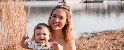 An adult with a light skin tone is sitting in front of a pond at sunset, holding a baby with a light skin tone, enjoying the peaceful setting.
