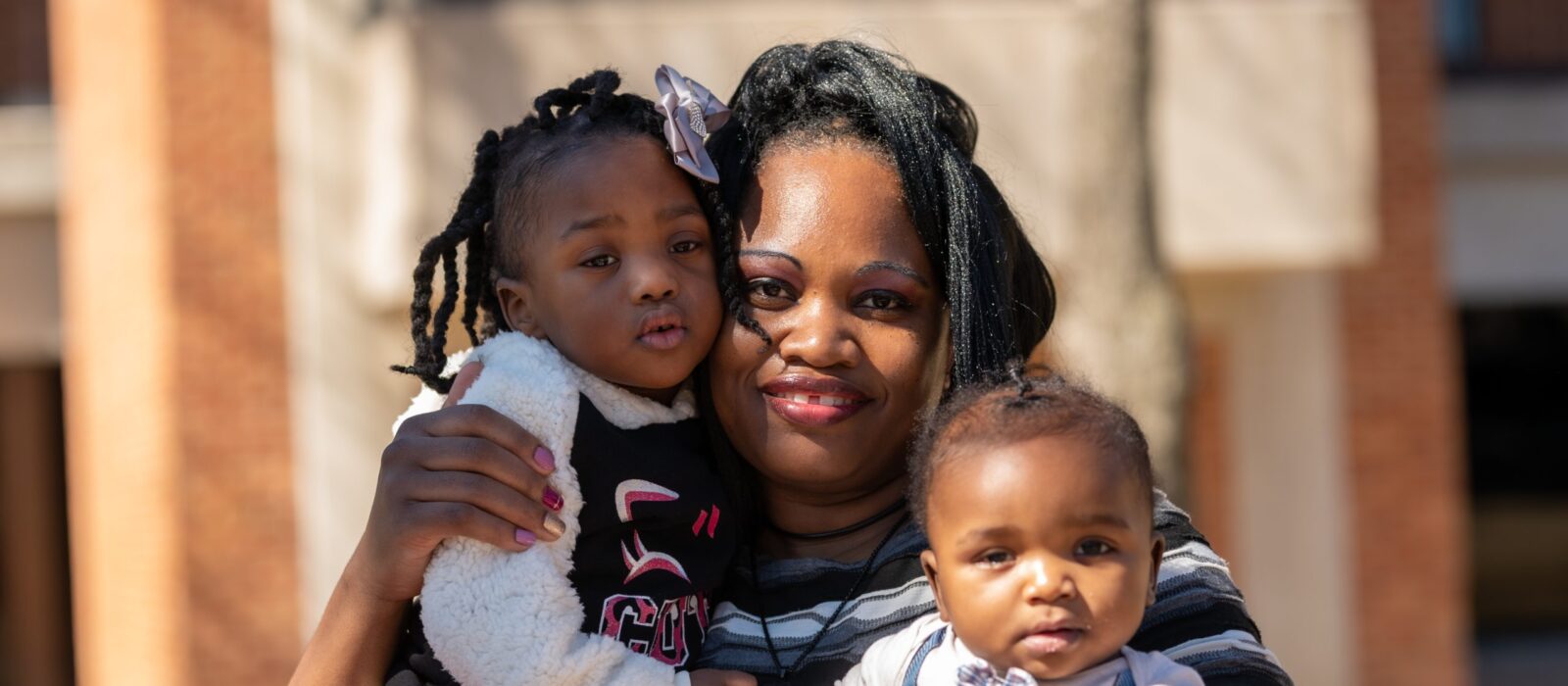 An adult with a dark skin tone is sitting on a bench with their baby with a dark skin tone on their lap and their young child with a dark skin tone standing beside them.